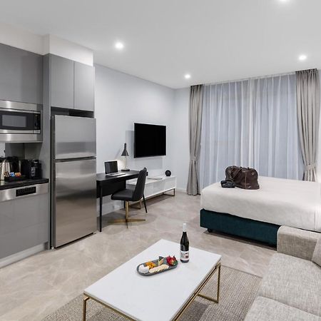 Meriton Suites Sussex Street, Σίδνεϊ Εξωτερικό φωτογραφία The photo shows a modern studio apartment. On one side, there is a kitchenette with sleek gray cabinets, a refrigerator, and built-in appliances. In the living area, there is a comfortable gray sofa, a white coffee table with a fruit platter and a bo