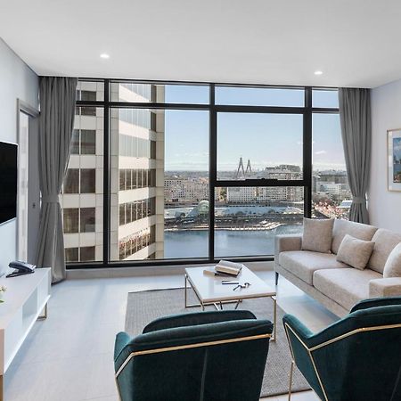 Meriton Suites Sussex Street, Σίδνεϊ Εξωτερικό φωτογραφία The photo shows a modern living room with large windows that offer a view of the cityscape and water. The interior features a light color palette with pale blue walls and soft lighting. There is a comfortable gray sofa and two green accent chairs, ar