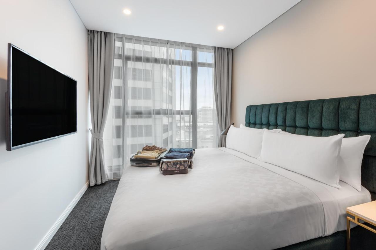Meriton Suites Sussex Street, Σίδνεϊ Εξωτερικό φωτογραφία The photo shows a modern hotel bedroom. The room features a large bed with a green upholstered headboard and crisp white linens. There are two pillows on the bed and a few folded items of clothing neatly placed on it. A flat-screen TV is mounted on t