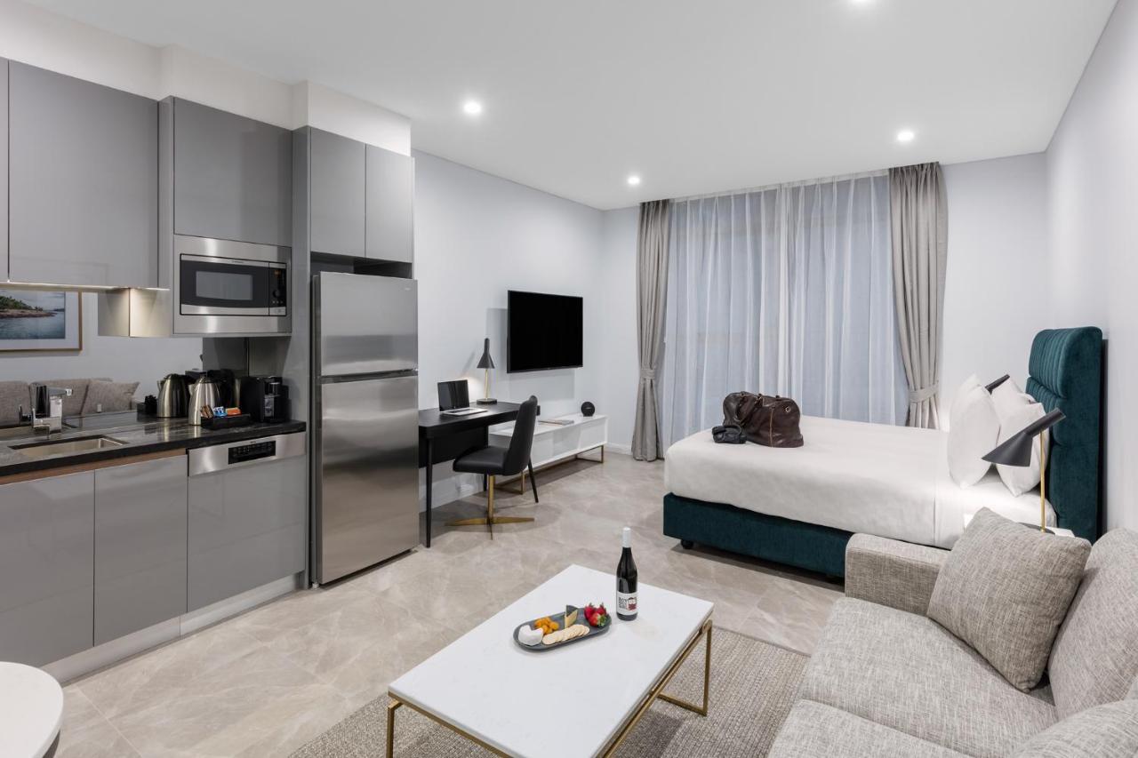 Meriton Suites Sussex Street, Σίδνεϊ Εξωτερικό φωτογραφία The photo shows a modern studio apartment. On one side, there is a kitchenette with sleek gray cabinets, a refrigerator, and built-in appliances. In the living area, there is a comfortable gray sofa, a white coffee table with a fruit platter and a bo