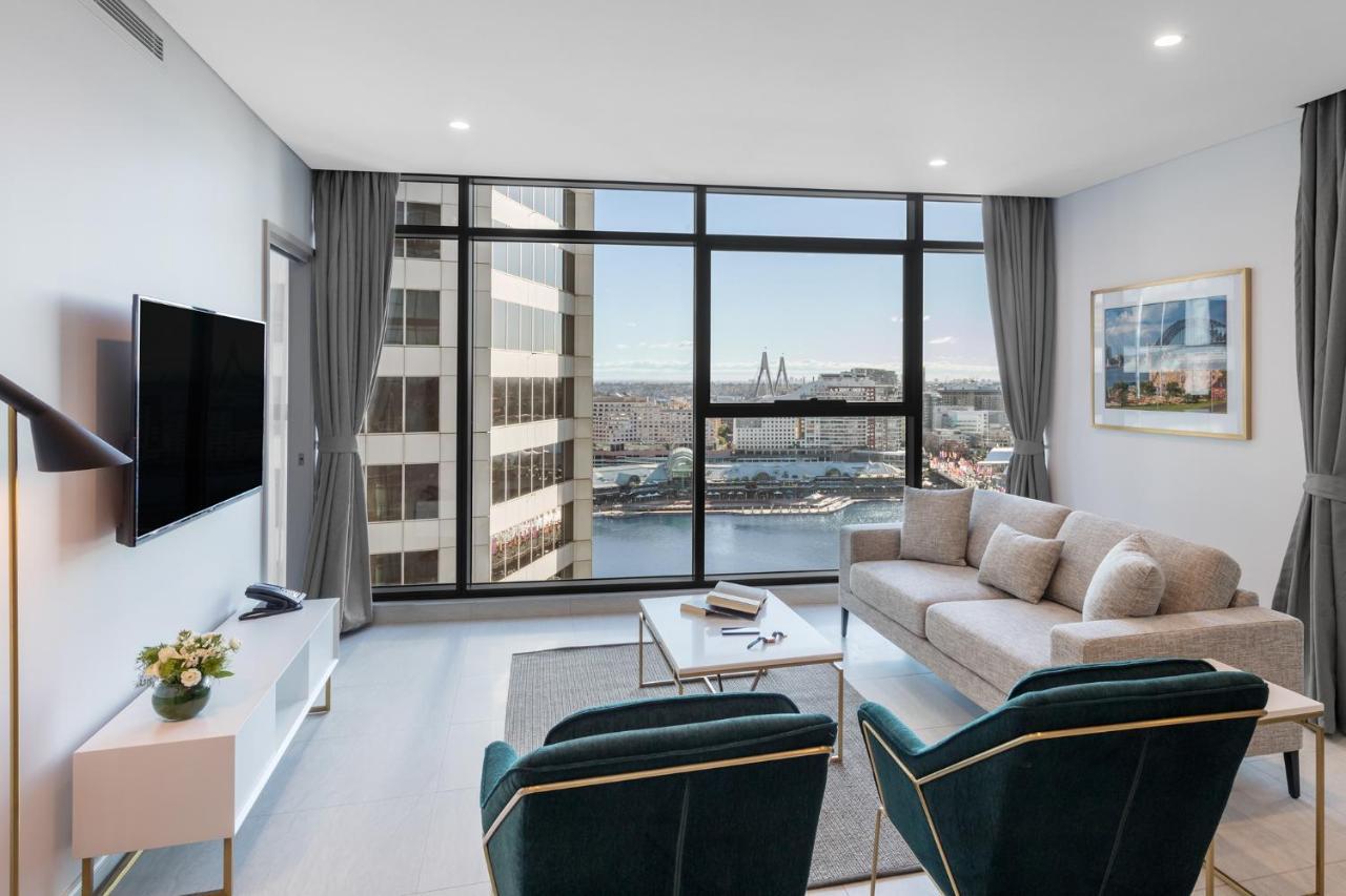 Meriton Suites Sussex Street, Σίδνεϊ Εξωτερικό φωτογραφία The photo shows a modern living room with large windows that offer a view of the cityscape and water. The interior features a light color palette with pale blue walls and soft lighting. There is a comfortable gray sofa and two green accent chairs, ar