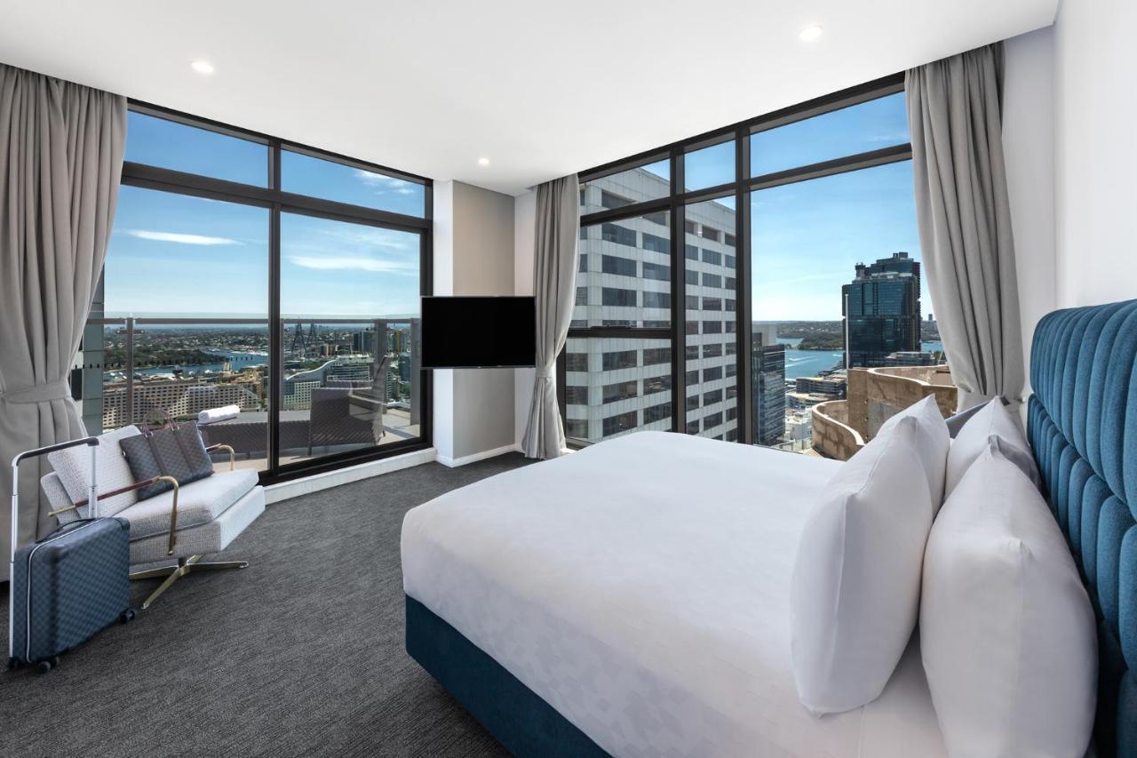 Meriton Suites Sussex Street, Σίδνεϊ Εξωτερικό φωτογραφία The photo shows a modern hotel room featuring large windows that provide a panoramic view of the cityscape and waterfront. The room is furnished with a king-size bed dressed in white linens, a comfortable armchair, and a flat-screen TV mounted on the