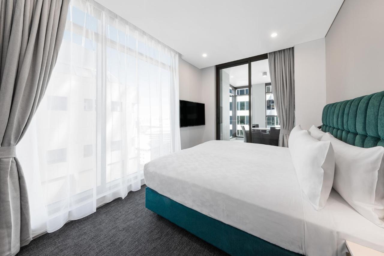 Meriton Suites Sussex Street, Σίδνεϊ Εξωτερικό φωτογραφία The photo shows a modern bedroom featuring a large bed with a white duvet and pillows. The headboard is turquoise, adding a pop of color to the neutral-toned room. There are sheer white curtains covering large windows that let in natural light, creat