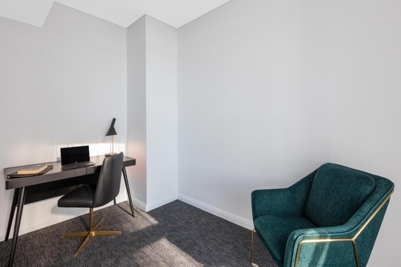 Meriton Suites Sussex Street, Σίδνεϊ Εξωτερικό φωτογραφία The photo depicts a minimalist interior space, likely a small office or study area. It features a simple, modern desk with a black lamp and a laptop or tablet on it. A sleek black chair accompanies the desk, while on the opposite side of the room, th
