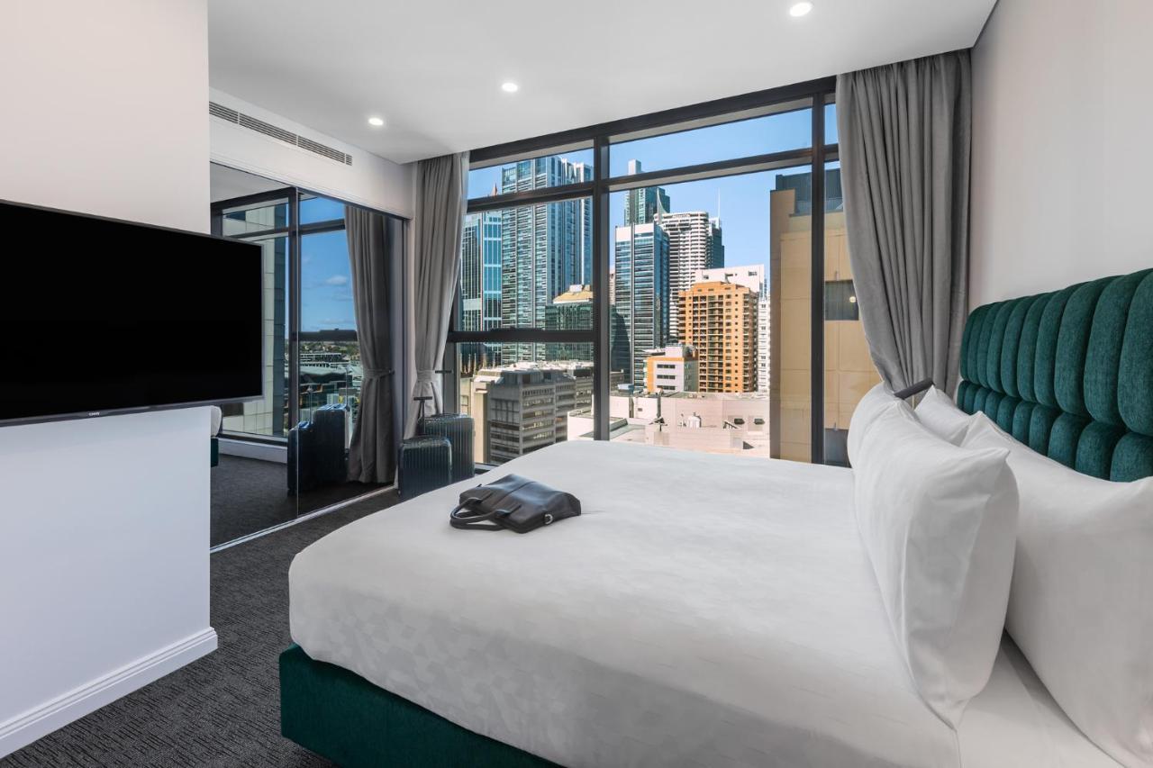 Meriton Suites Sussex Street, Σίδνεϊ Εξωτερικό φωτογραφία The photo shows a modern hotel room featuring a large bed with white linens and a green upholstered headboard. There is a sleek television mounted on the wall opposite the bed, and the room has large windows that provide a view of a cityscape, includ