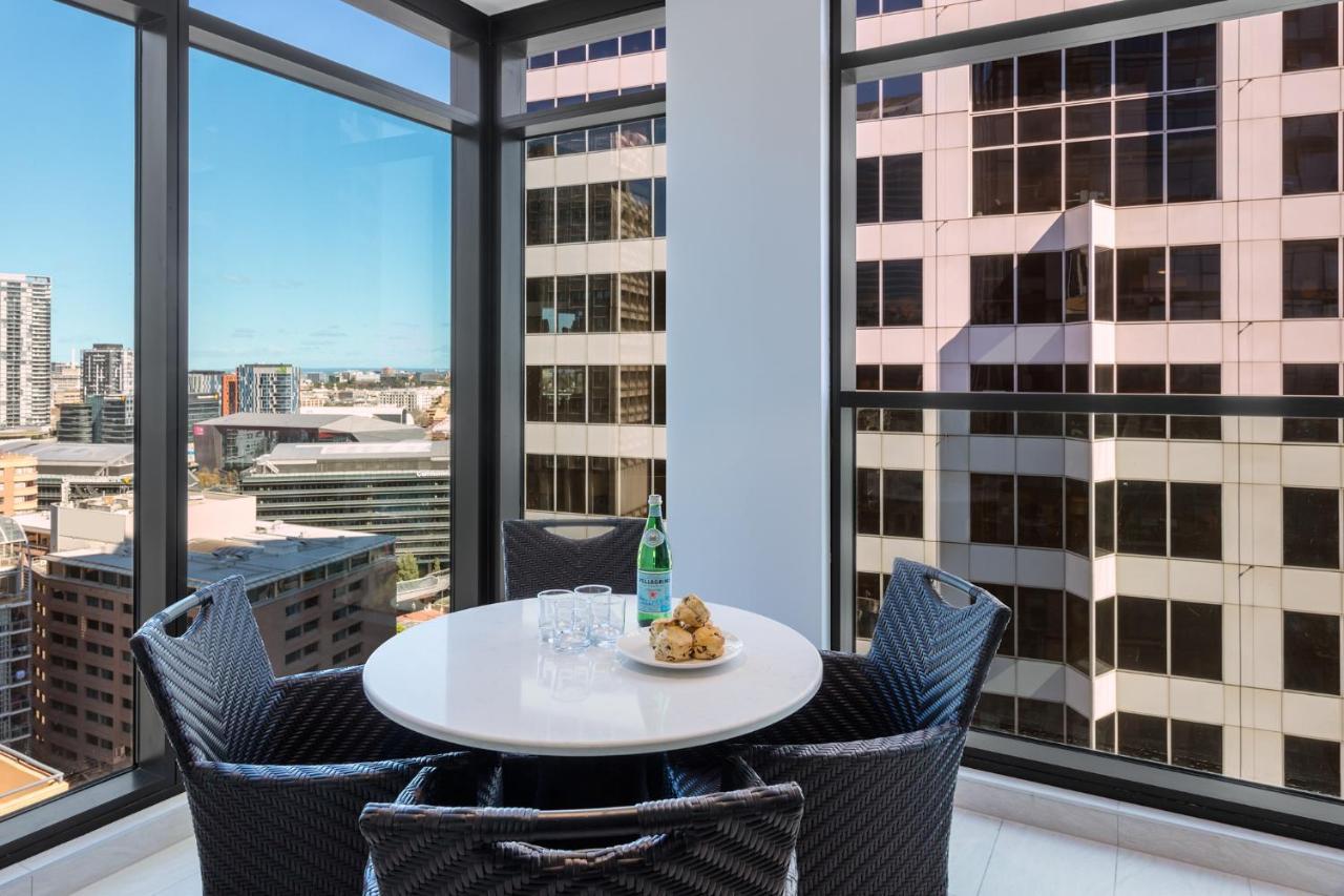 Meriton Suites Sussex Street, Σίδνεϊ Εξωτερικό φωτογραφία The photo shows a modern dining space with a round white table set for four. There are four black woven chairs around the table. On the table, there's a bottle of sparkling water, two glasses, and a small plate with several cookies. The large windows
