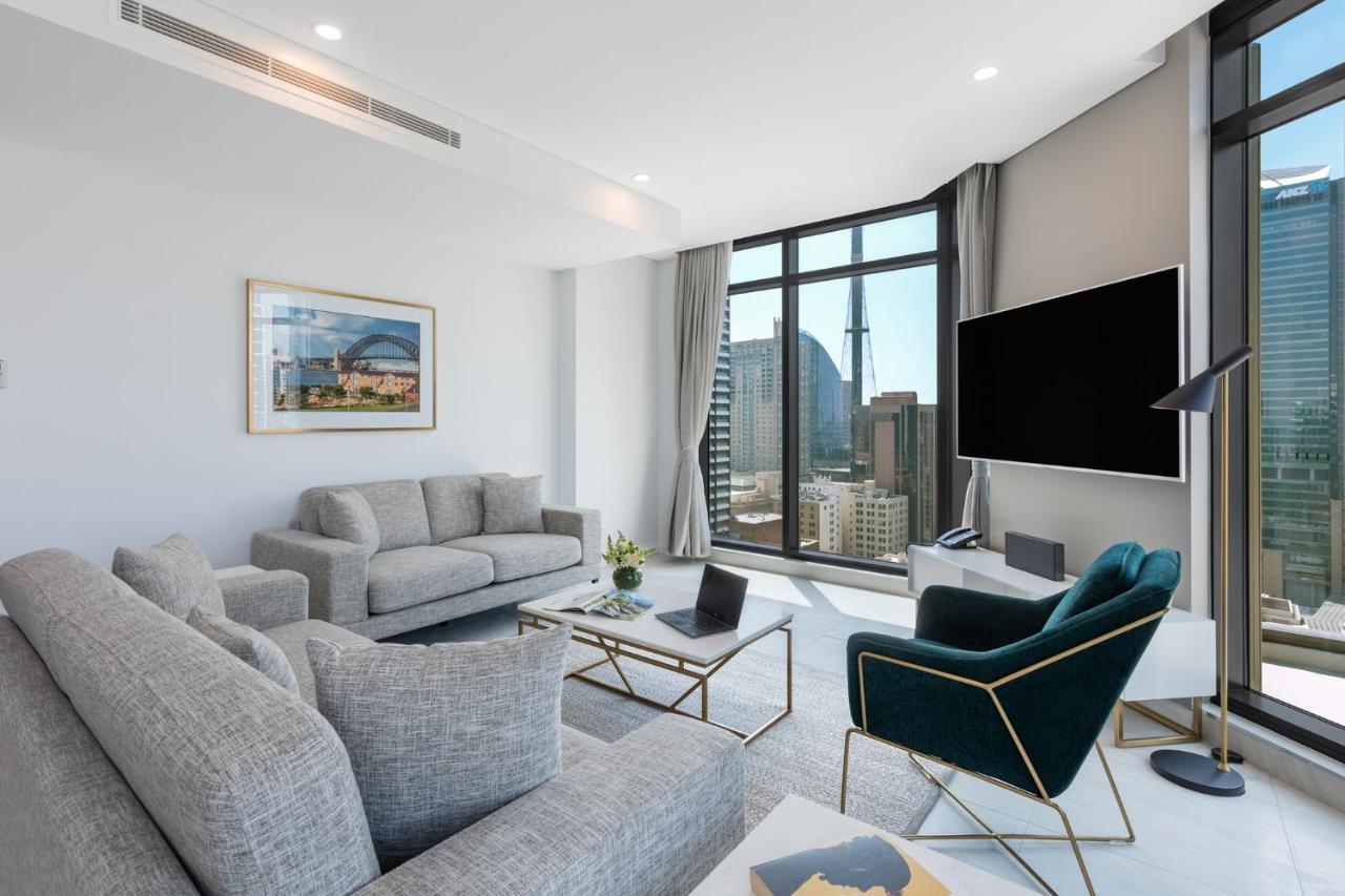 Meriton Suites Sussex Street, Σίδνεϊ Εξωτερικό φωτογραφία The photo shows a modern living room with large windows offering a view of a city skyline. The room features two gray sofas and a stylish green accent chair. A sleek coffee table is placed in front of the sofas, alongside a laptop. There’s a flat-scr