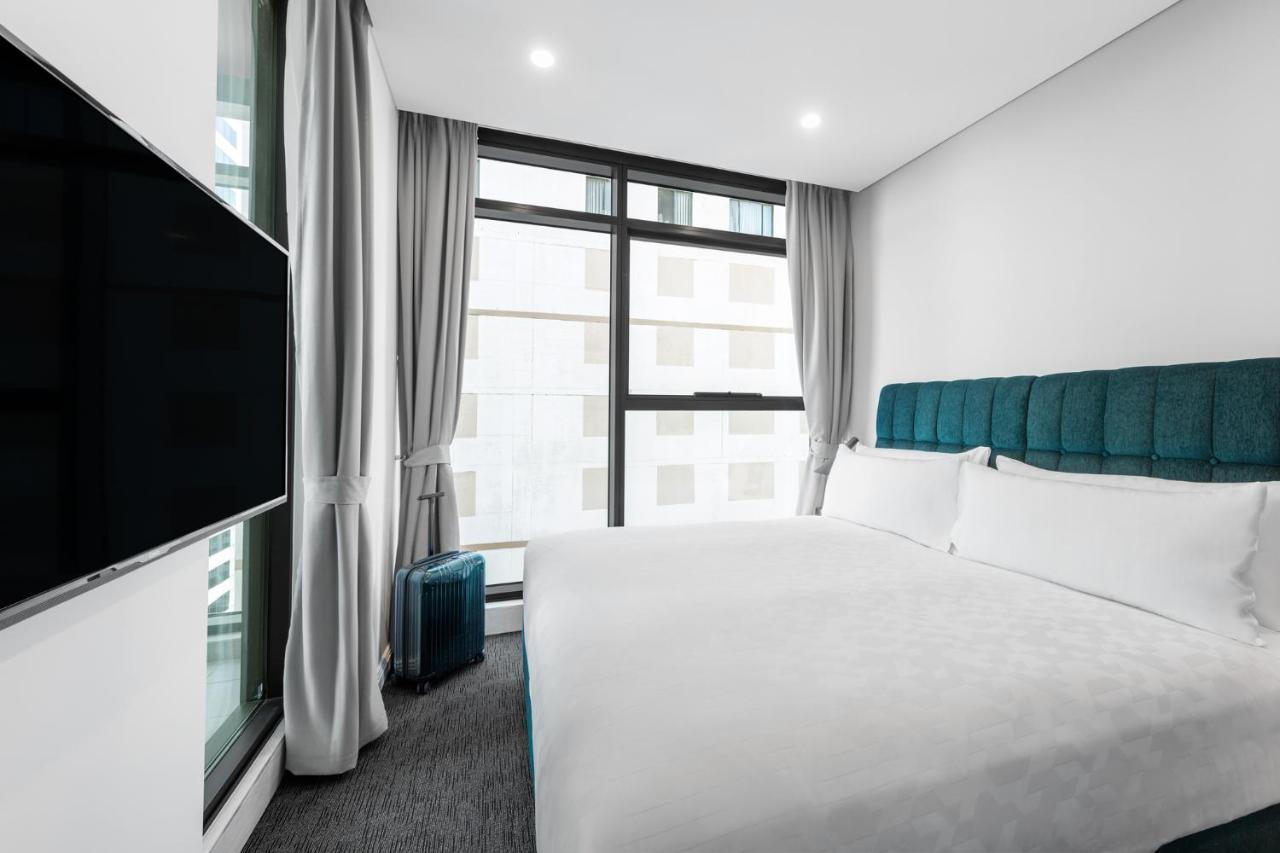Meriton Suites Sussex Street, Σίδνεϊ Εξωτερικό φωτογραφία The photo shows a modern hotel room featuring a large bed with a light-colored duvet and a blue upholstered headboard. There are two pillows on the bed. A large window allows natural light to enter the room, and gray curtains frame the window. Next t