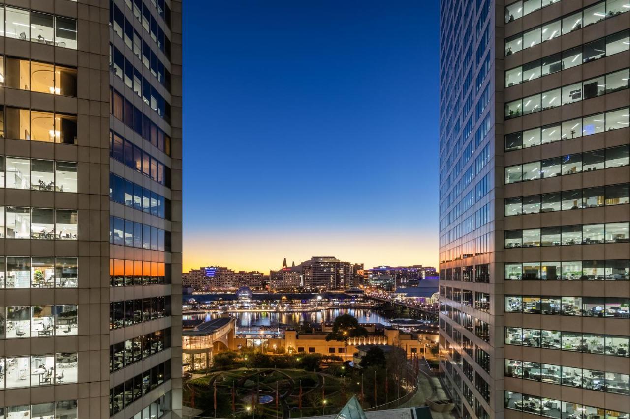 Meriton Suites Sussex Street, Σίδνεϊ Εξωτερικό φωτογραφία The photo shows a view between two tall buildings, likely office towers, during twilight. On one side, there are glass windows reflecting the evening sky that transitions from blue to orange near the horizon. In the background, a cityscape is visible