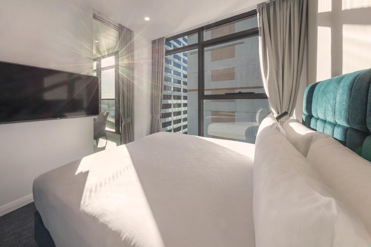 Meriton Suites Sussex Street, Σίδνεϊ Εξωτερικό φωτογραφία The photo shows a modern hotel room with a neatly made bed featuring white bedding and a plush, tufted headboard in a shade of blue. Natural light is streaming through large windows, which are covered partially by light gray curtains. A flat-screen t