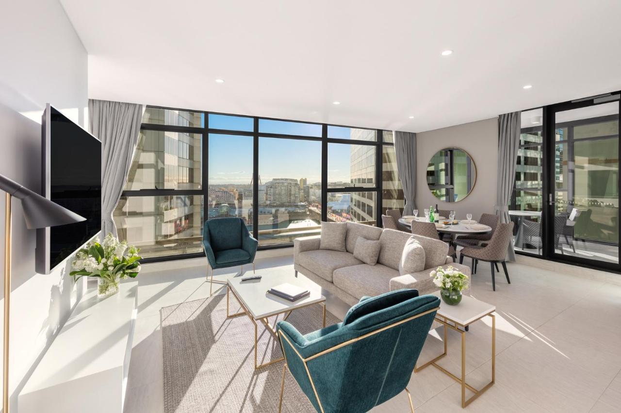Meriton Suites Sussex Street, Σίδνεϊ Εξωτερικό φωτογραφία The photo shows a modern and stylish living space. It features a spacious room with large windows that provide a bright view of the cityscape. The furniture includes a light gray sofa, two teal chairs, and a coffee table. There is a flat-screen TV mo