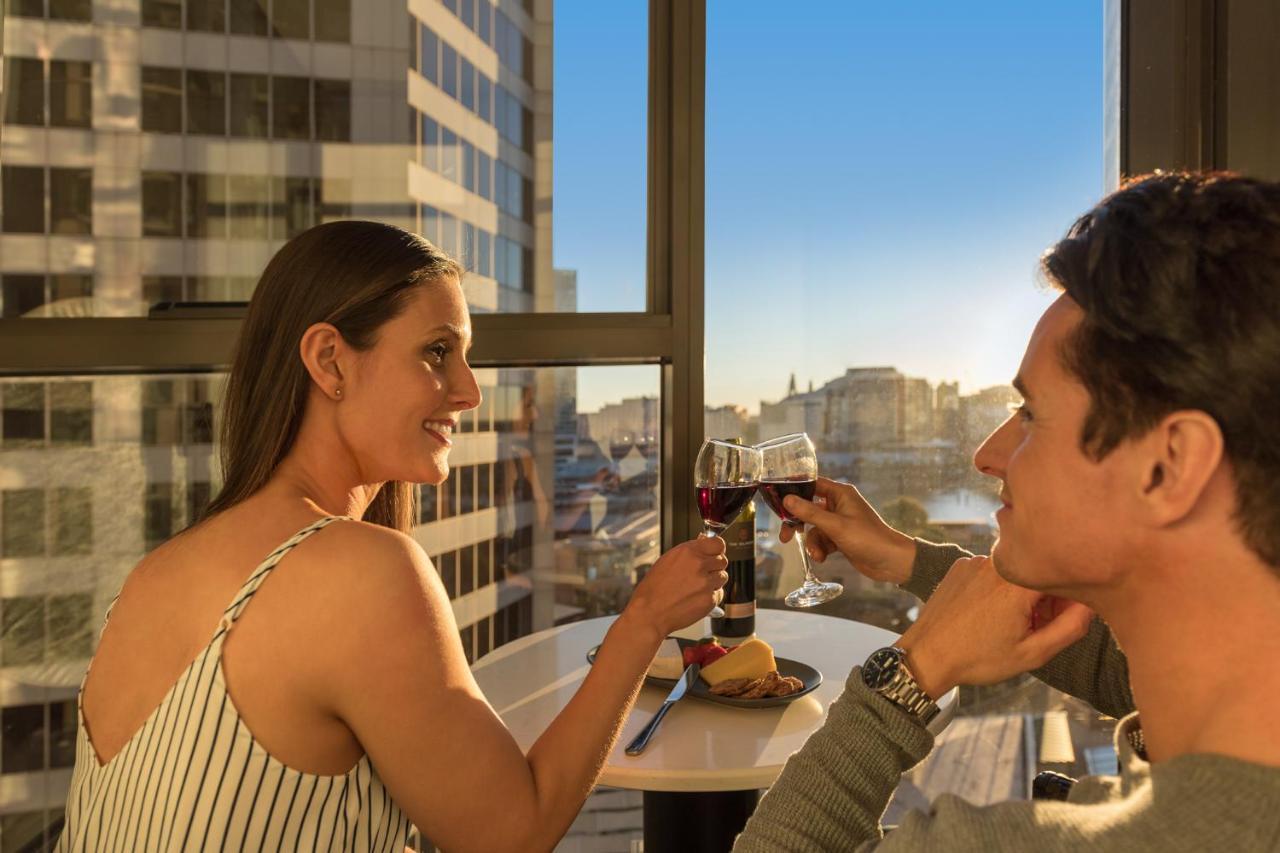 Meriton Suites Sussex Street, Σίδνεϊ Εξωτερικό φωτογραφία The photo depicts a couple enjoying a drink together in a modern setting. They are sitting at a small table near a large window, which provides a view of a cityscape and a sunset. Both individuals are smiling and engaged in a cheerful toast with glas