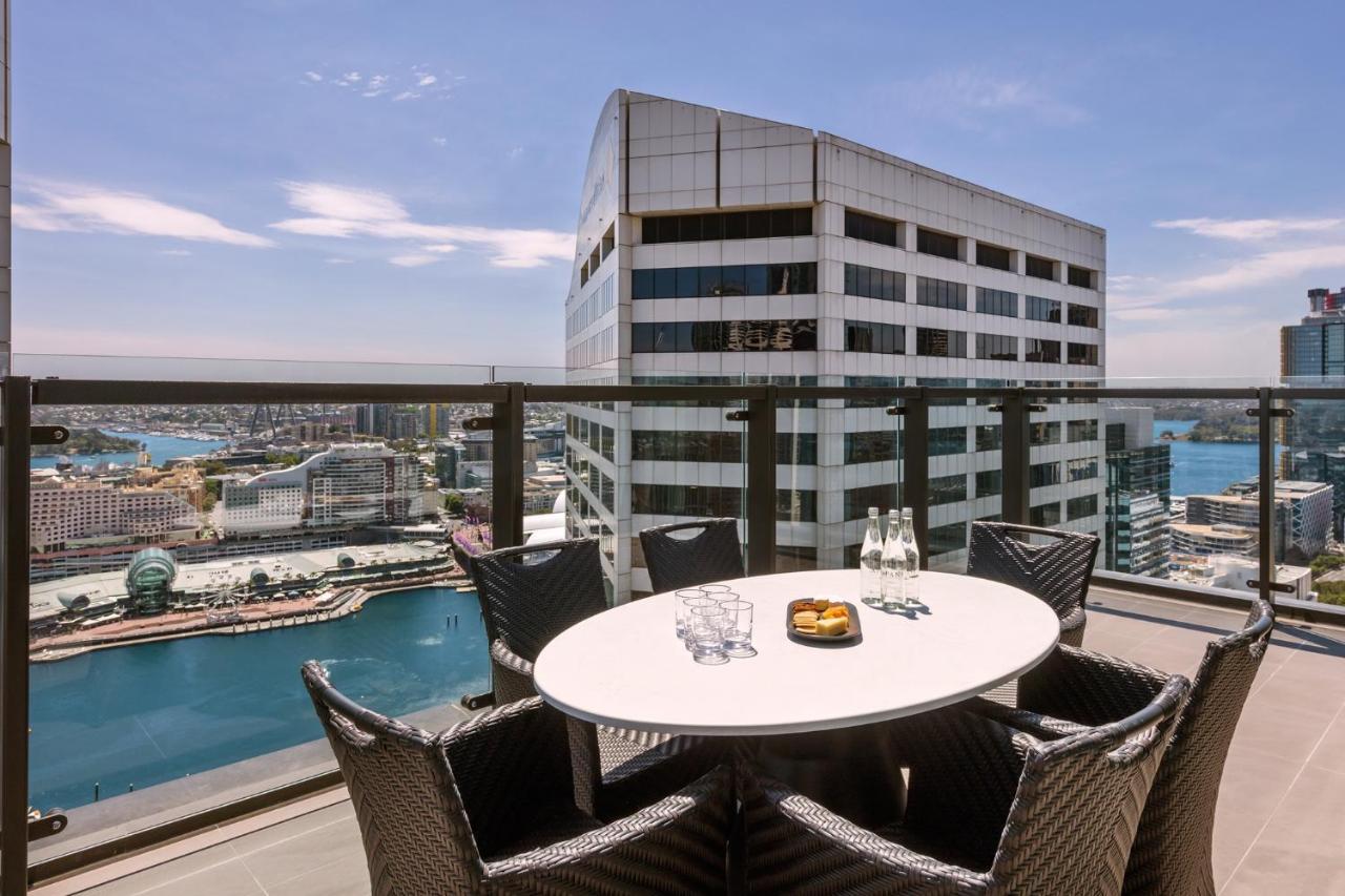 Meriton Suites Sussex Street, Σίδνεϊ Εξωτερικό φωτογραφία The photo shows a balcony or terrace with a round table and several chairs arranged around it. The table is set with some drinks and a plate, possibly containing food. In the background, there is a view of a waterway and several buildings, including 