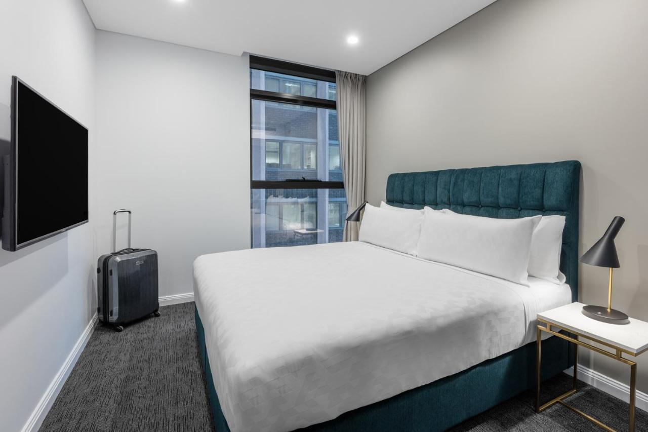 Meriton Suites Sussex Street, Σίδνεϊ Εξωτερικό φωτογραφία The photo shows a modern hotel room with a clean and minimalist design. There is a large, neatly made bed with white bedding and a teal-colored headboard. On one side of the bed, there is a small nightstand with a lamp. A sleek television is mounted 