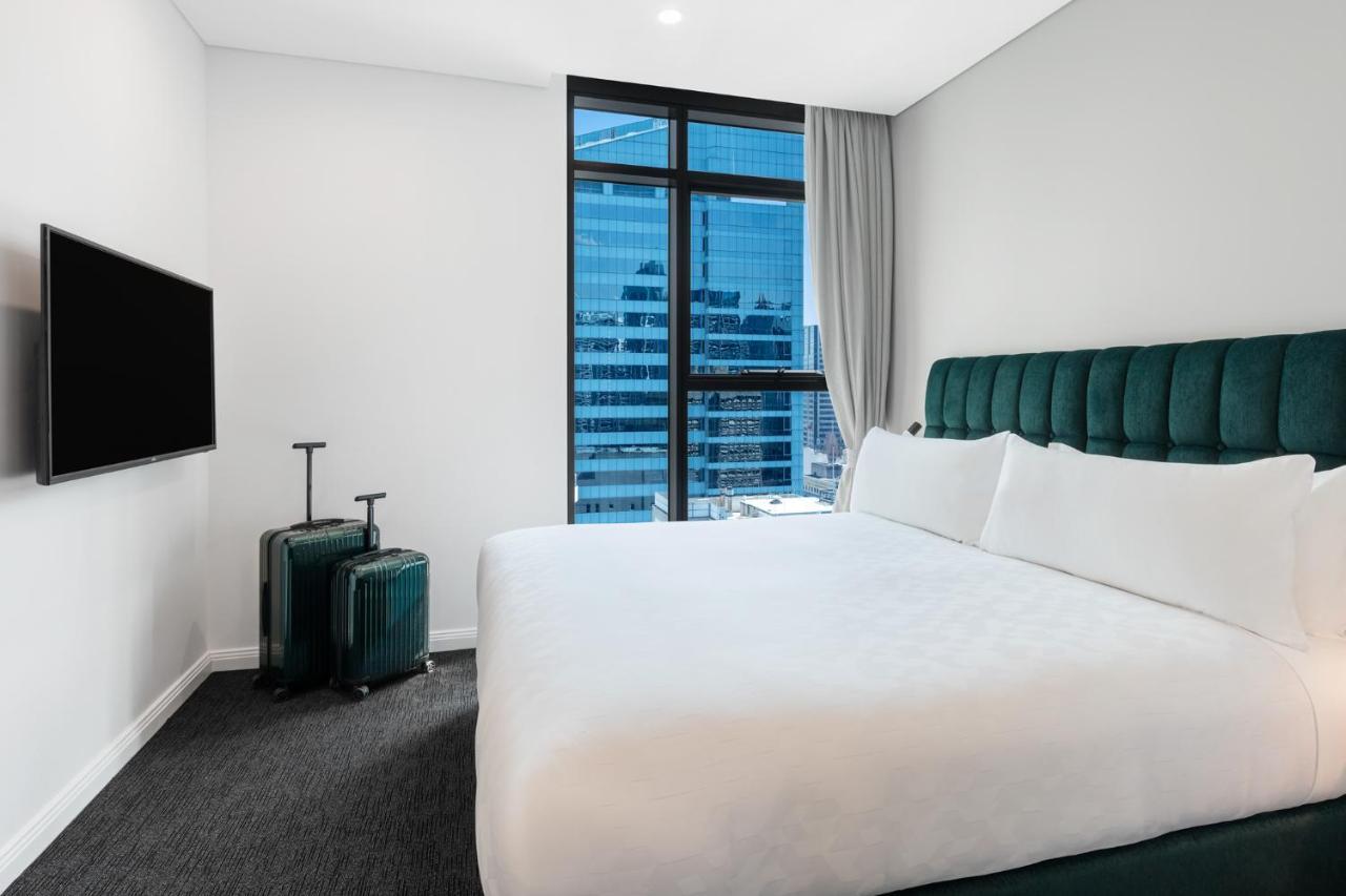 Meriton Suites Sussex Street, Σίδνεϊ Εξωτερικό φωτογραφία The photo shows a modern hotel room featuring a large bed with white bedding and a green upholstered headboard. There are two green suitcases placed beside the bed. A flat-screen TV is mounted on the wall opposite the bed. The room has a large window