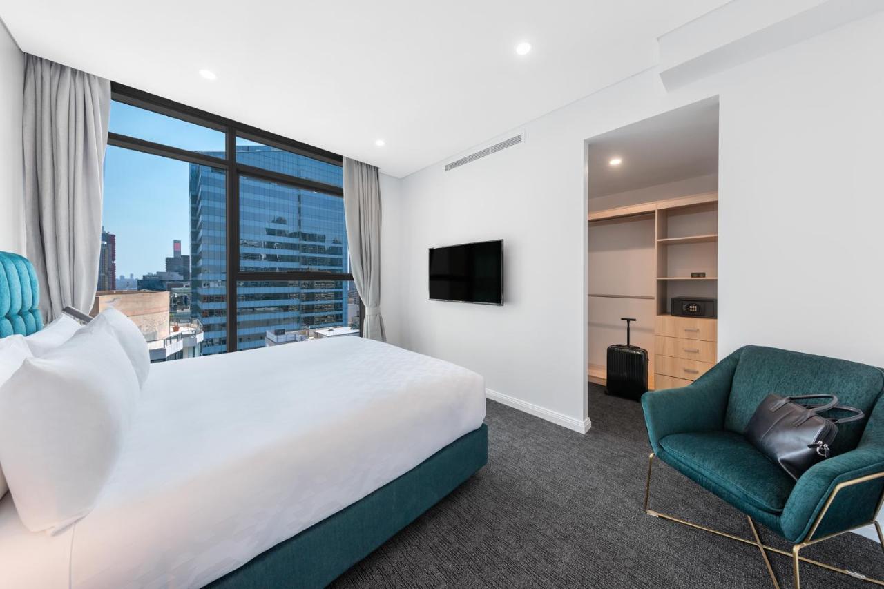 Meriton Suites Sussex Street, Σίδνεϊ Εξωτερικό φωτογραφία The photo shows a modern hotel room. It features a large window that allows natural light to fill the space, offering a view of the city skyline. The room includes a neatly made bed with a white duvet and a teal-colored base. There is a small green a