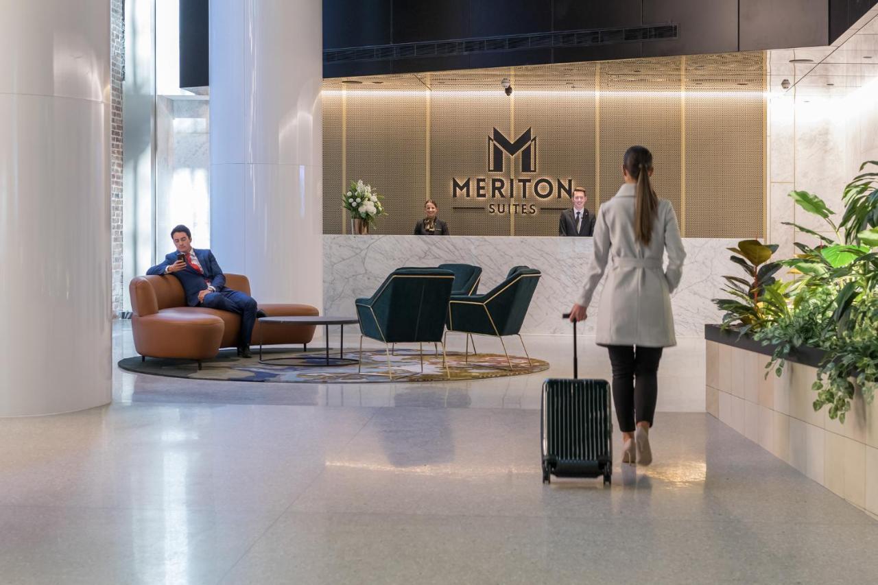Meriton Suites Sussex Street, Σίδνεϊ Εξωτερικό φωτογραφία The photo depicts a modern hotel lobby. In the foreground, a woman is walking towards the reception desk, pulling a suitcase behind her. She is dressed in a light-colored coat. To her left, there’s a comfortable seating area with a person sitting on 