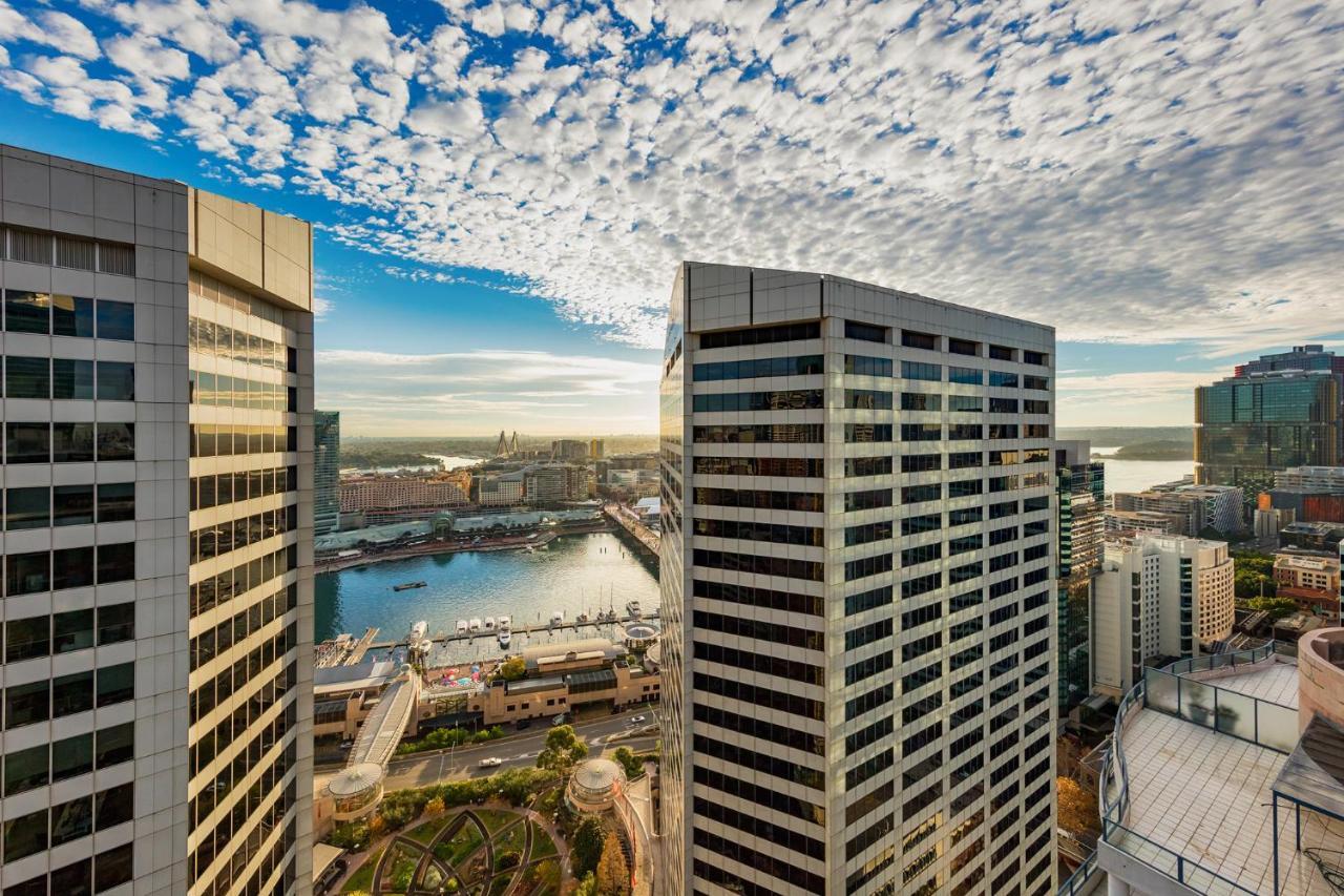 Meriton Suites Sussex Street, Σίδνεϊ Εξωτερικό φωτογραφία The photo depicts a panoramic view of a modern cityscape featuring tall commercial buildings with glass facades. The shot is taken from a height, showing the intricate architecture of the nearby structures. Below, there is a body of water, which appe