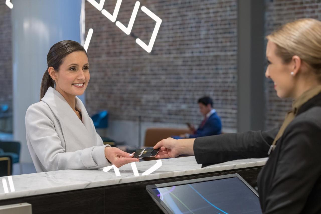 Meriton Suites Sussex Street, Σίδνεϊ Εξωτερικό φωτογραφία The image depicts a scene at a hotel reception desk. A woman with long hair, dressed in a white outfit, is smiling as she hands her card to a receptionist, who is in a black uniform. The reception area features a modern design, and there is a person 