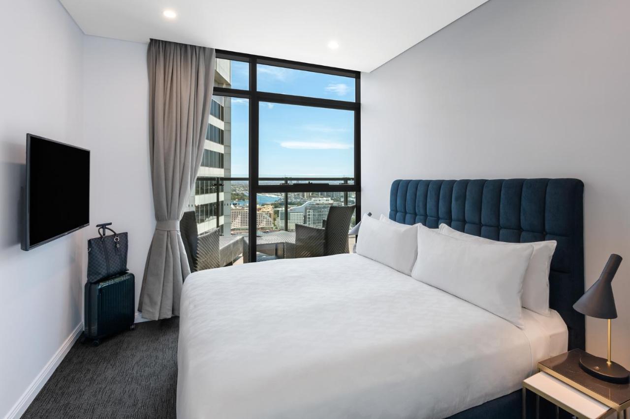 Meriton Suites Sussex Street, Σίδνεϊ Εξωτερικό φωτογραφία The photo shows a modern hotel room featuring a double bed with white linens and a tufted blue headboard. There is a small bedside table next to the bed. A large window allows natural light to fill the room, offering a view of the city outside, inclu