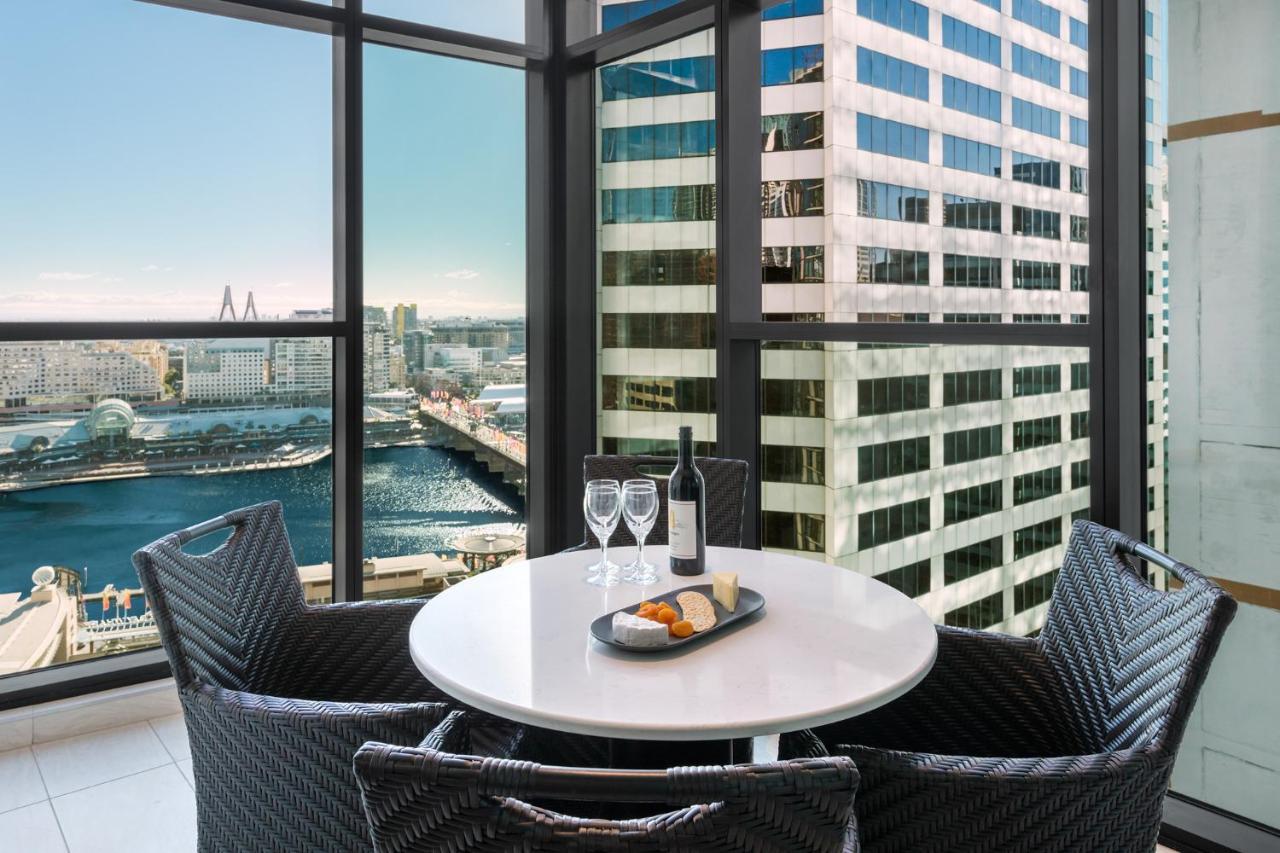 Meriton Suites Sussex Street, Σίδνεϊ Εξωτερικό φωτογραφία The photo shows a modern dining area with a round table and four woven chairs. On the table, there are two wine glasses, a bottle of wine, and a plate with food items. The large windows provide a view of a cityscape featuring tall buildings and a bod