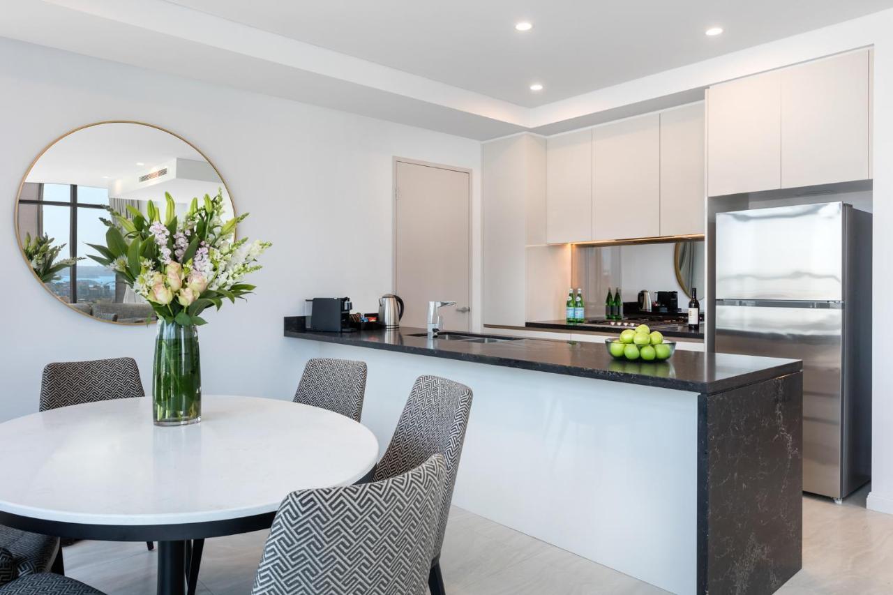 Meriton Suites Sussex Street, Σίδνεϊ Εξωτερικό φωτογραφία The photo shows a modern kitchen and dining area. In the foreground, there's a circular dining table surrounded by four stylish chairs. A large vase filled with fresh flowers is placed on the table, adding a touch of color and elegance. 

In the kitc