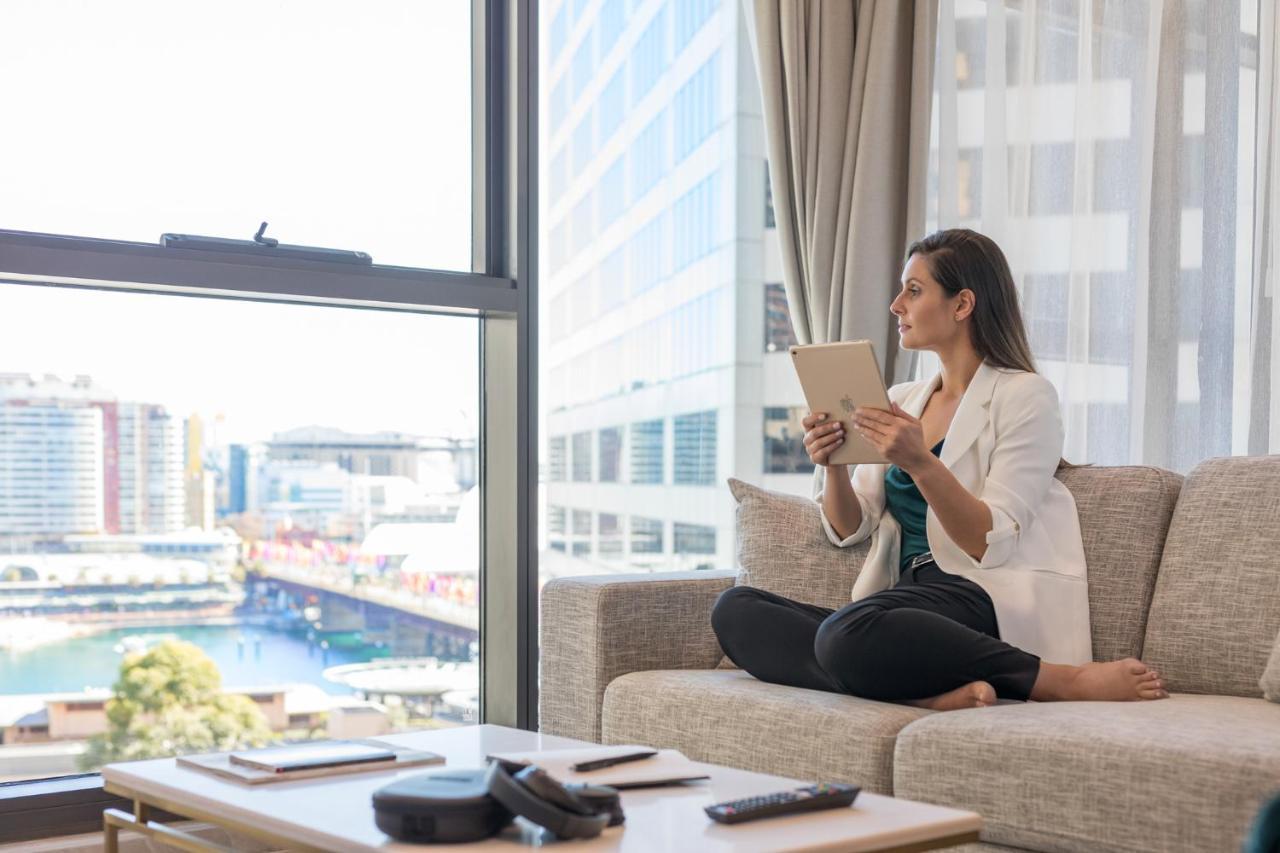 Meriton Suites Sussex Street, Σίδνεϊ Εξωτερικό φωτογραφία The image portrays a woman sitting cross-legged on a light-colored sofa, engaged with a tablet in her hands. She is dressed in a white blazer and dark pants, appearing relaxed and focused. The setting features large windows that provide a view of a w