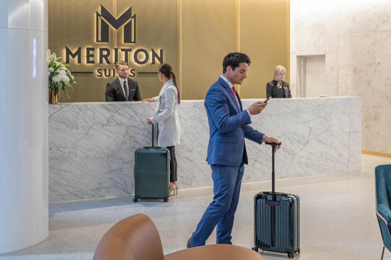 Meriton Suites Sussex Street, Σίδνεϊ Εξωτερικό φωτογραφία The photo shows the interior of a hotel reception area, specifically at Meriton Suites. In the foreground, a man in a blue suit is standing with a suitcase, looking at his phone. Behind him, there are two staff members at the reception desk, one of w