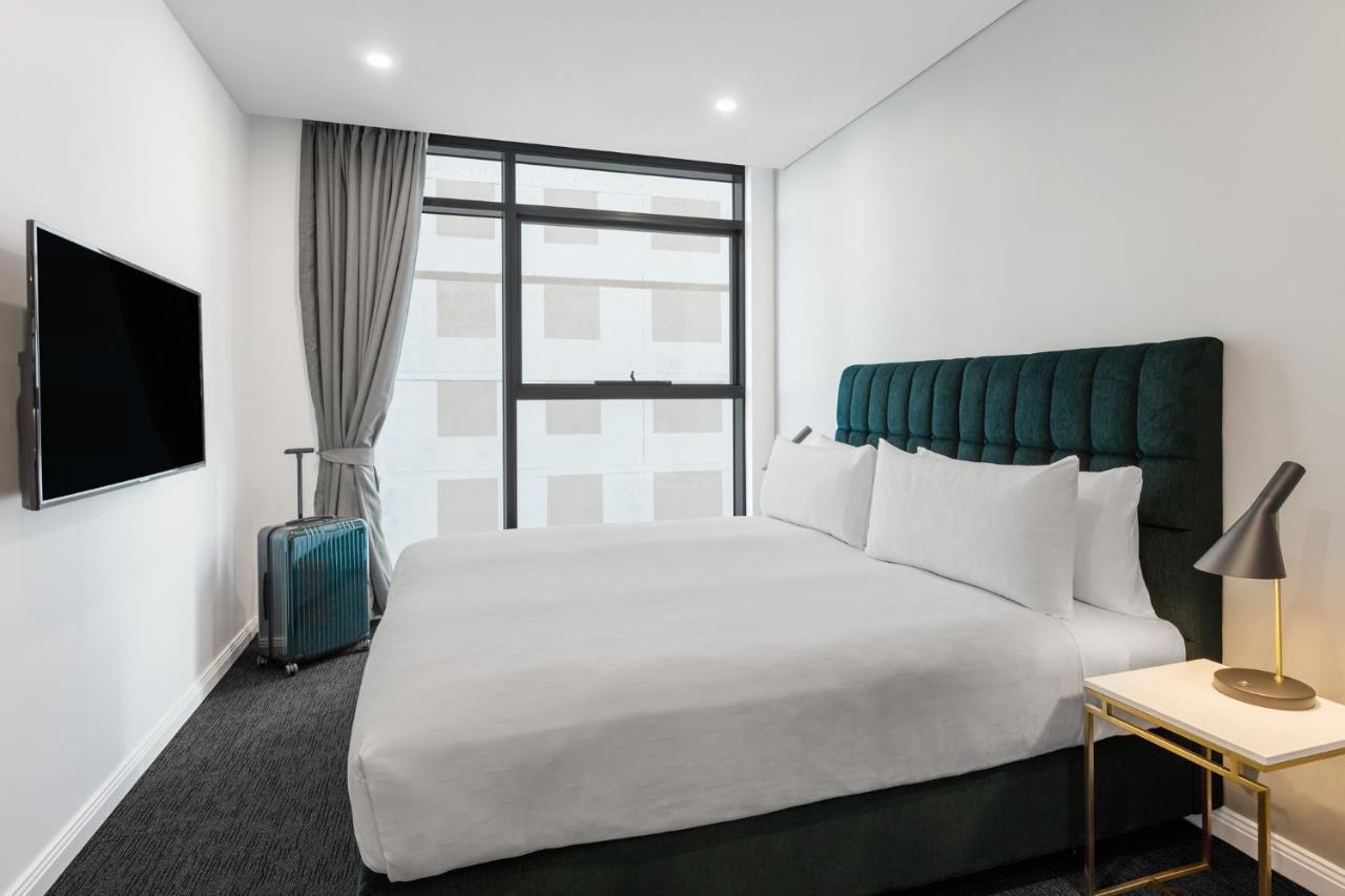 Meriton Suites Sussex Street, Σίδνεϊ Εξωτερικό φωτογραφία The photo shows a modern hotel room. It features a large bed with a dark green tufted headboard and multiple white pillows. There's a nightstand with a lamp beside the bed. A sleek television is mounted on the wall opposite the bed, and a suitcase is
