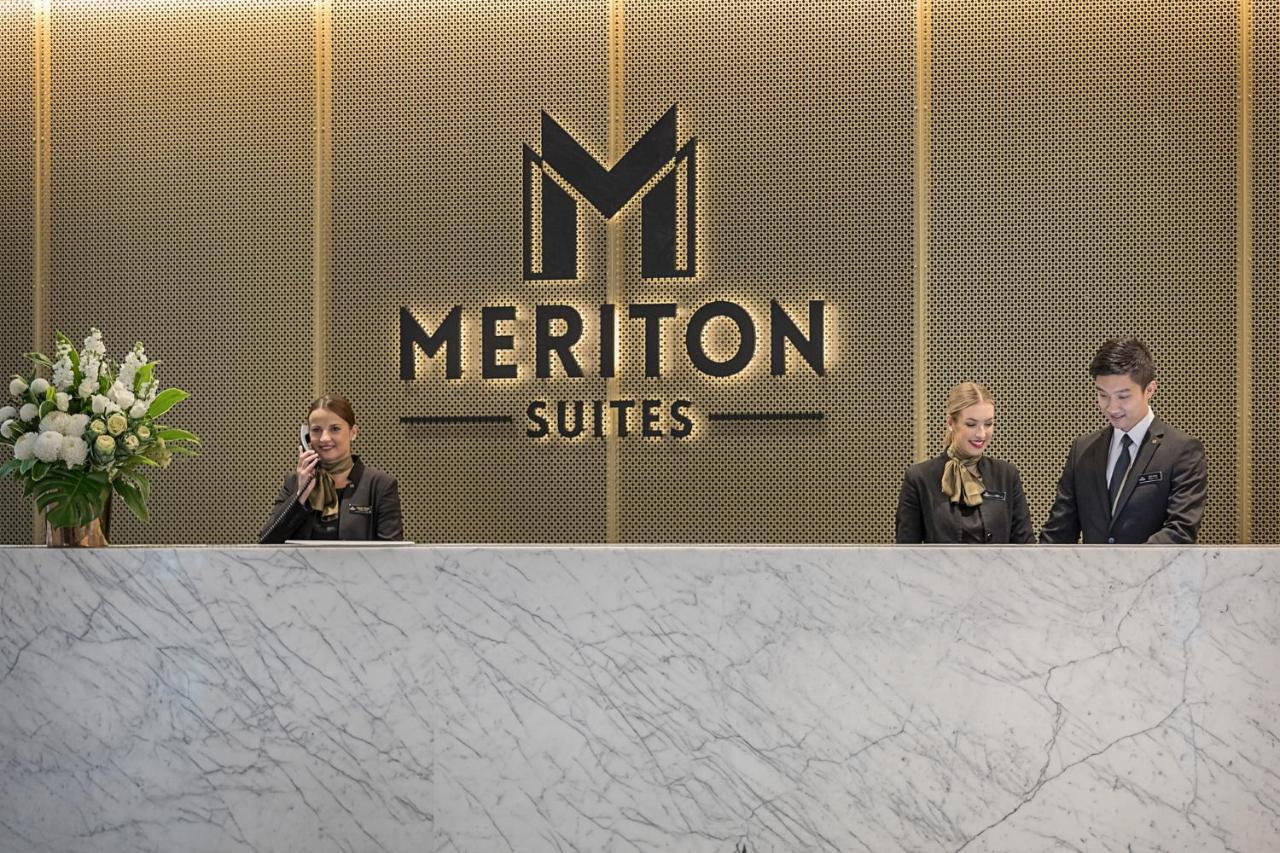 Meriton Suites Sussex Street, Σίδνεϊ Εξωτερικό φωτογραφία The photo depicts the reception area of a hotel called "Meriton Suites." In the background, there is a large, illuminated sign featuring the hotel’s logo. The reception desk is made of marble and is staffed by two individuals dressed in professional 
