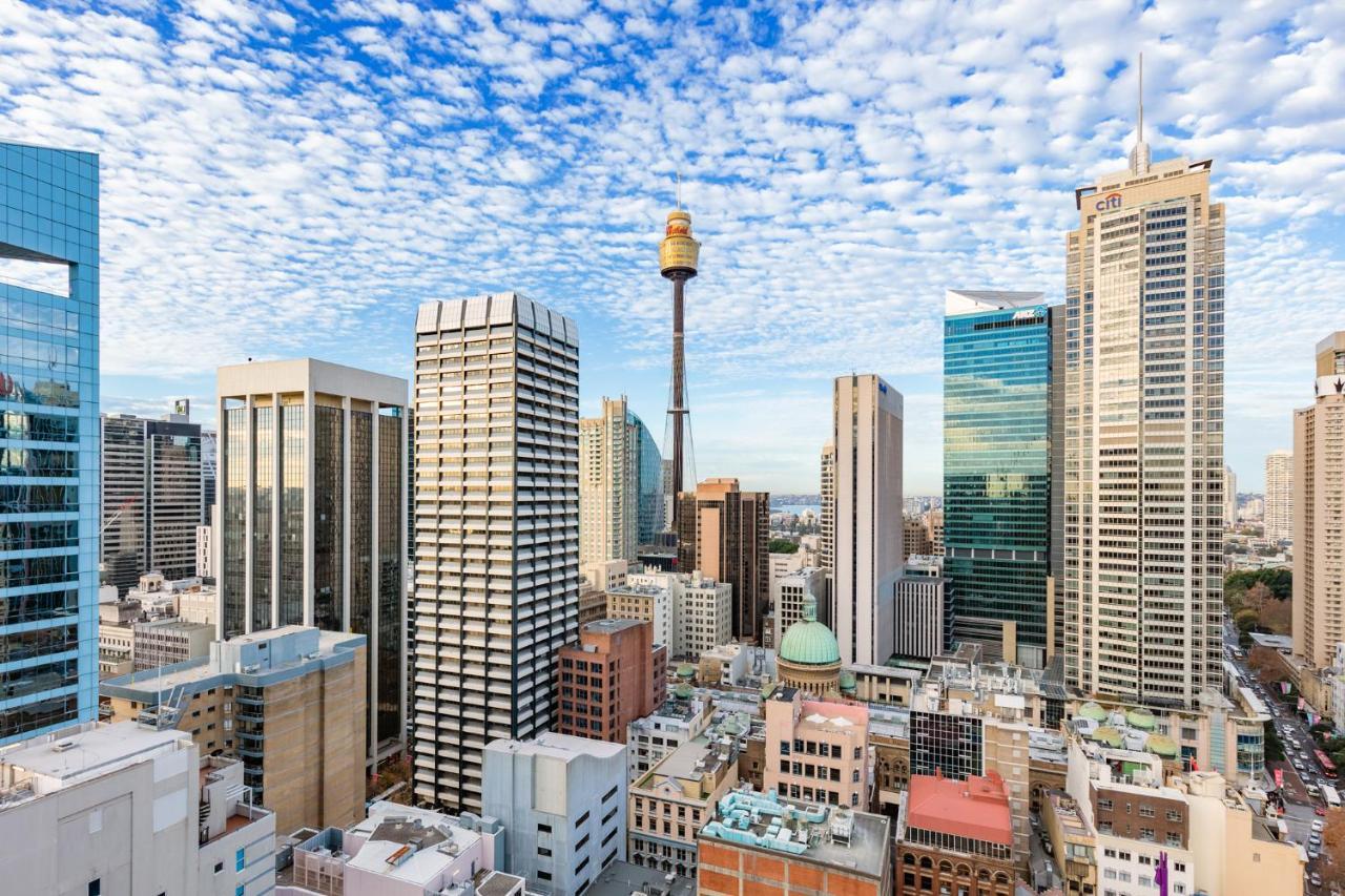 Meriton Suites Sussex Street, Σίδνεϊ Εξωτερικό φωτογραφία The photo shows a panoramic view of a city skyline dominated by tall skyscrapers. In the center, there is a notable tower with a distinctive golden structure at its top, which is likely a landmark of the city. The buildings have a mix of modern glass
