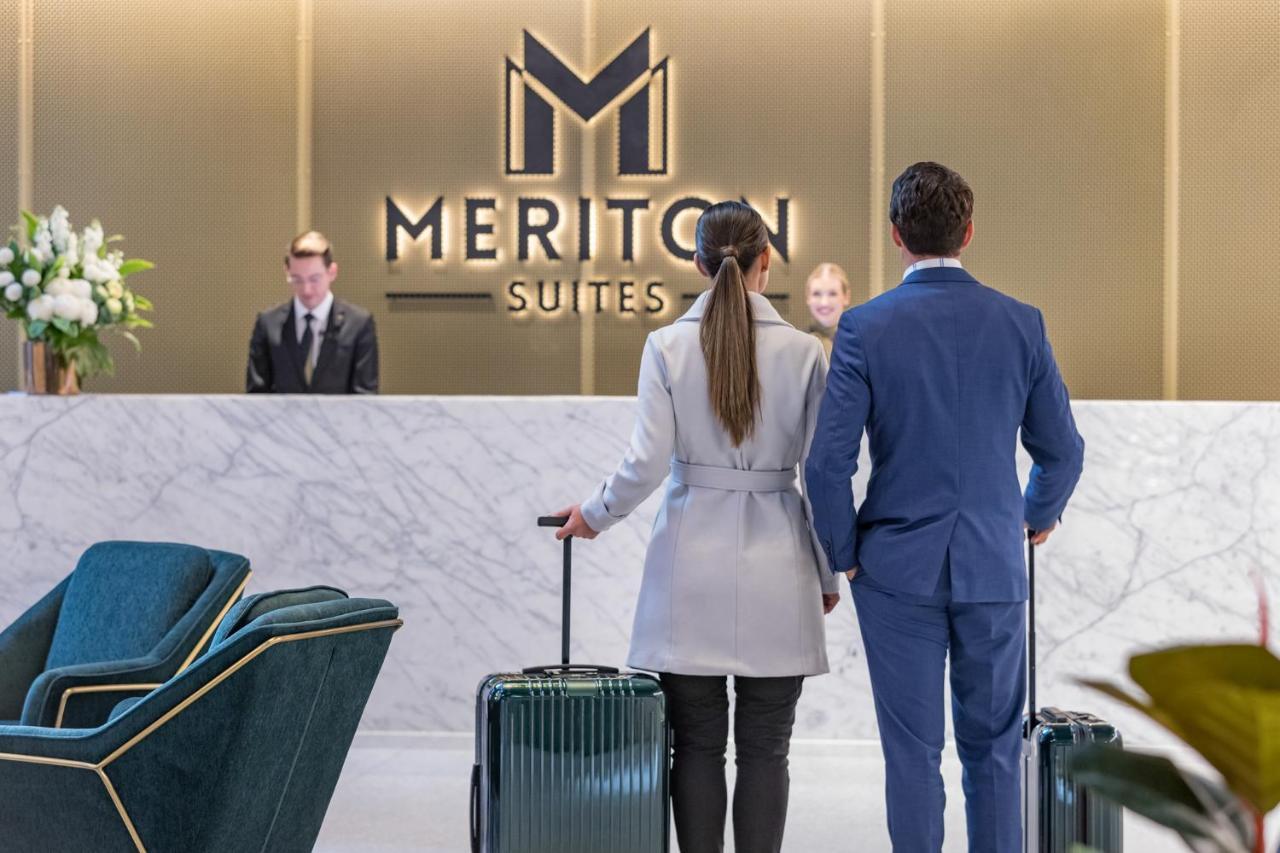 Meriton Suites Sussex Street, Σίδνεϊ Εξωτερικό φωτογραφία The photo shows a couple standing at a reception area in a hotel named "Meriton Suites." They are both facing the reception desk, where staff members are visible. The man is dressed in a suit, and the woman is wearing a light-colored coat. They each 