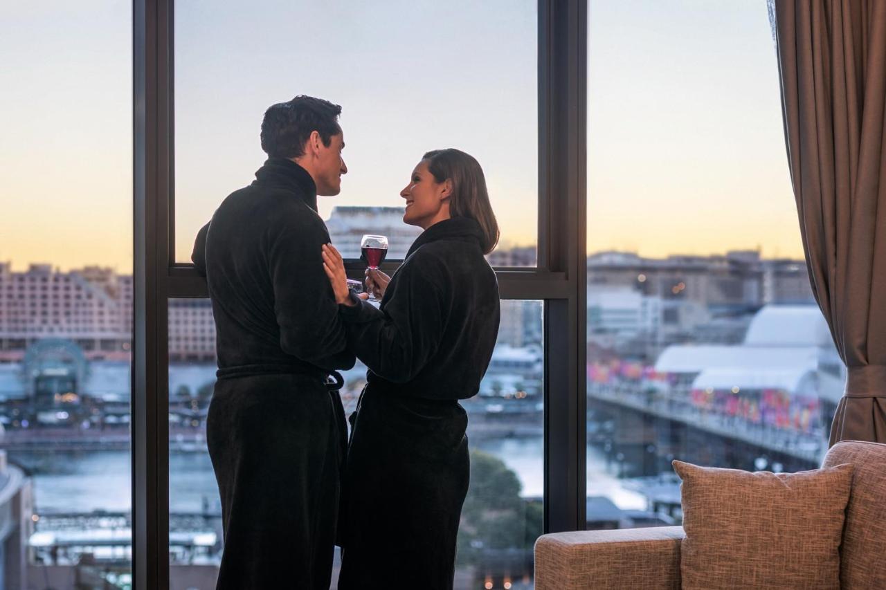 Meriton Suites Sussex Street, Σίδνεϊ Εξωτερικό φωτογραφία The photo depicts a couple standing closely together by a large window. They are wearing dark bathrobes and holding drinks, gazing into each other’s eyes. The background features a scenic view, likely of a cityscape at sunset, with soft light illumin