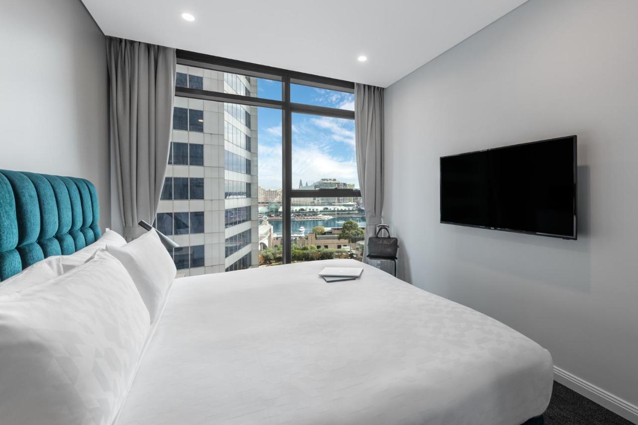 Meriton Suites Sussex Street, Σίδνεϊ Εξωτερικό φωτογραφία The photo shows a modern hotel room with a large bed featuring a white comforter and pillows. There is a blue headboard that adds a pop of color to the decor. A large window provides a view of the outside, which includes buildings and a glimpse of wa