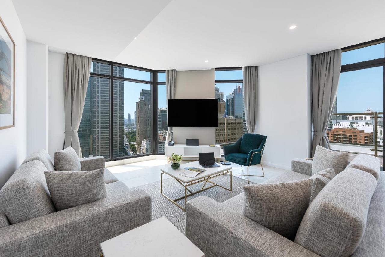 Meriton Suites Sussex Street, Σίδνεϊ Εξωτερικό φωτογραφία The photo showcases a modern living room with large windows offering a view of a city skyline. The room features a light color palette with gray sofas arranged around a coffee table. A flat-screen TV is mounted on the wall, and there is a green armch