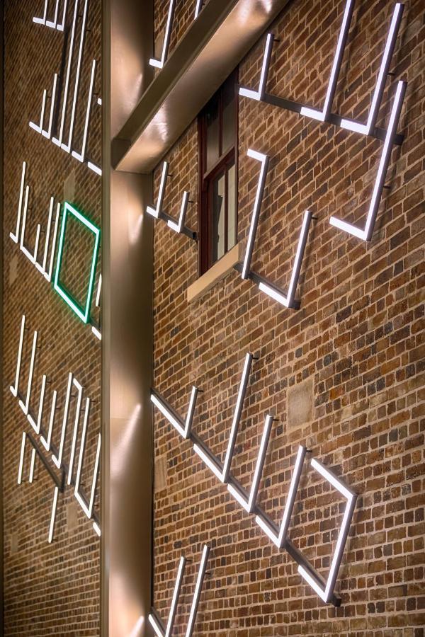 Meriton Suites Sussex Street, Σίδνεϊ Εξωτερικό φωτογραφία The photo shows a brick wall featuring a design made up of linear light fixtures. These light strips create geometric shapes and patterns, with some sections illuminated in white and one section highlighted in green. The combination of the brick text