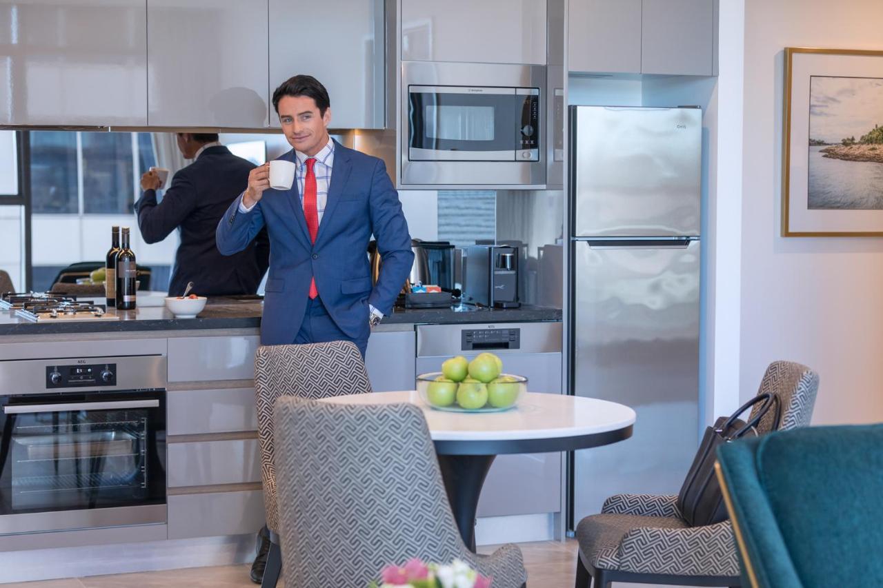 Meriton Suites Sussex Street, Σίδνεϊ Εξωτερικό φωτογραφία The photo depicts a well-dressed man in a suit standing in a modern kitchen. He is holding a cup, possibly of coffee, and appears to be looking towards the camera. The kitchen features contemporary appliances, including a microwave and refrigerator. 